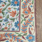 Corner view of the Gulmarg rug featuring intricate floral embroidery with vivid blue, orange, and green threads bordered by a cream background on wooden flooring.