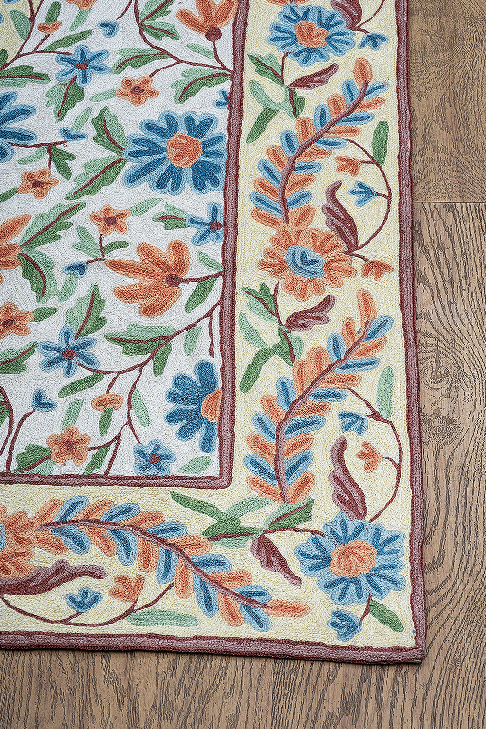 Corner view of the Gulmarg rug featuring intricate floral embroidery with vivid blue, orange, and green threads bordered by a cream background on wooden flooring.