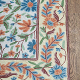 Corner view of the Gulmarg rug featuring intricate floral embroidery with vivid blue, orange, and green threads bordered by a cream background on wooden flooring.