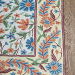 Corner view of the Gulmarg rug featuring intricate floral embroidery with vivid blue, orange, and green threads bordered by a cream background on wooden flooring.