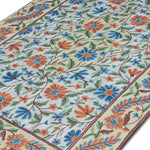Close-up angled view of the Gulmarg floral rug highlighting intricate chain stitch embroidery and vibrant blue, orange, and green floral patterns on a cream background.