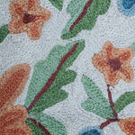 Extreme close-up of the Gulmarg rug showing detailed chain stitch embroidery with textured orange flowers, green leaves, and blue buds on a cream wool background.