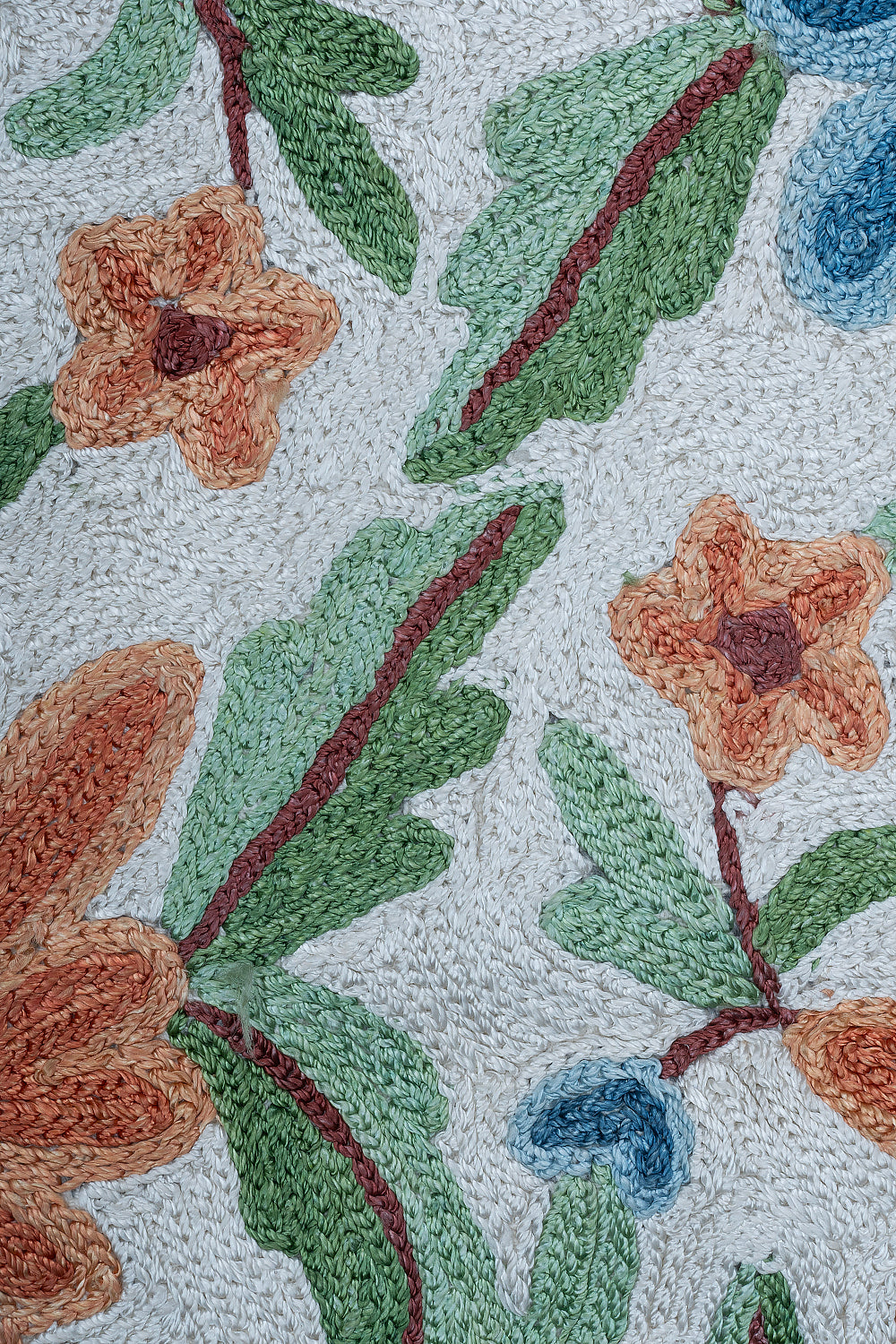 Extreme close-up of the Gulmarg rug showing detailed chain stitch embroidery with textured orange flowers, green leaves, and blue buds on a cream wool background.