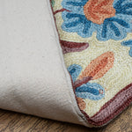 Close-up of the Gulmarg rug corner flipped to reveal the durable cotton backing. The visible floral embroidery on the front showcases textured blue and orange blossoms with maroon outlining on a cream base, resting on a wooden floor.