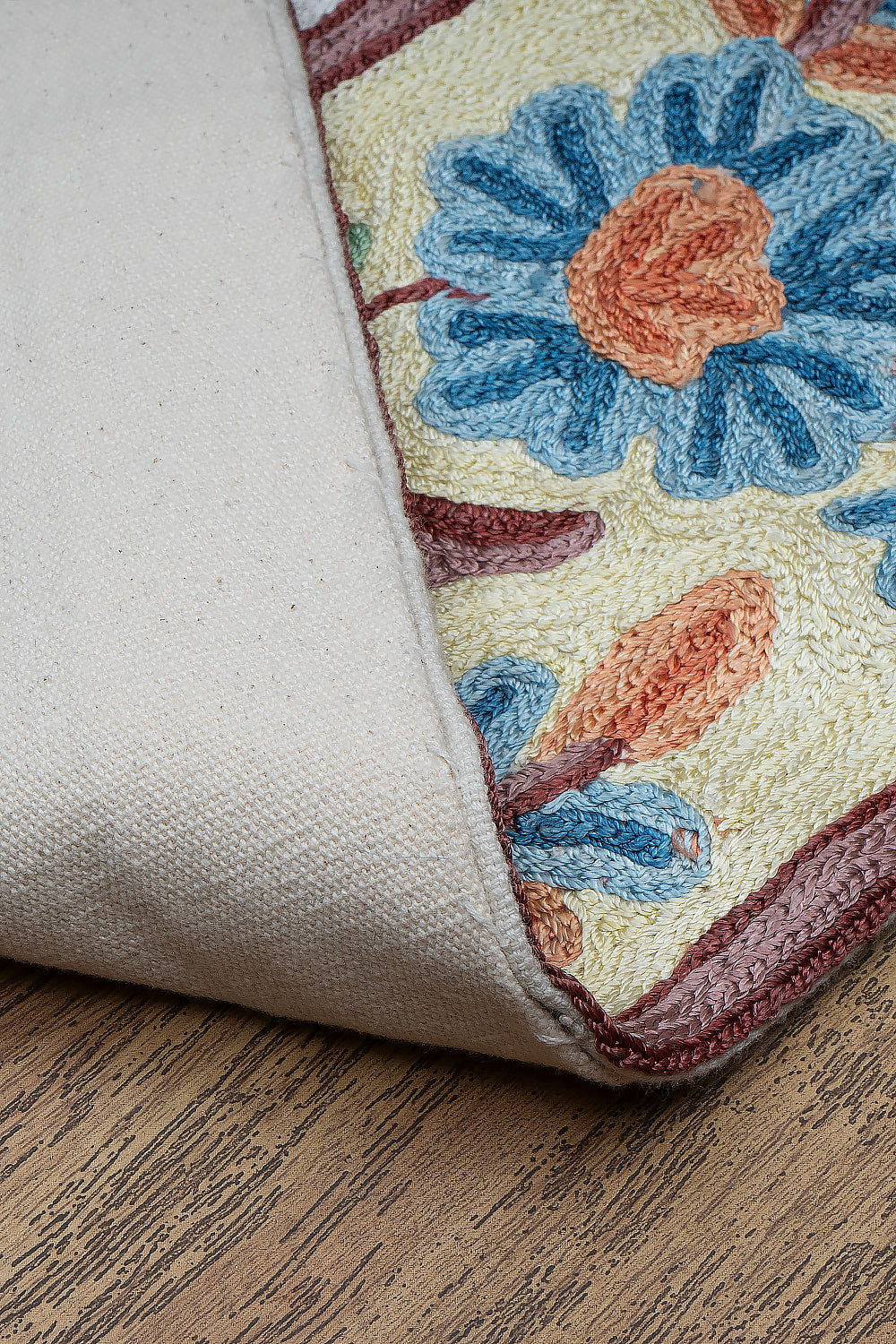 Close-up of the Gulmarg rug corner flipped to reveal the durable cotton backing. The visible floral embroidery on the front showcases textured blue and orange blossoms with maroon outlining on a cream base, resting on a wooden floor.