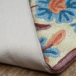 Close-up of the Gulmarg rug corner flipped to reveal the durable cotton backing. The visible floral embroidery on the front showcases textured blue and orange blossoms with maroon outlining on a cream base, resting on a wooden floor.