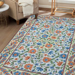 Handcrafted Gulmarg floral rug placed in a cozy living room, featuring intricate chain stitch embroidery in cream, blue, orange, and green tones complementing the neutral interior décor.