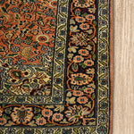 A close-up of the corner of a Kirman Persian rug, highlighting its intricate floral border design and delicate fringe. The rich peach and navy hues contrast beautifully with the finely detailed green and ivory patterns. The rug is placed on a light wooden floor, emphasizing its elegant craftsmanship.