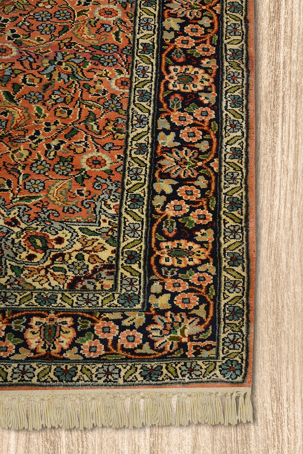 A close-up of the corner of a Kirman Persian rug, highlighting its intricate floral border design and delicate fringe. The rich peach and navy hues contrast beautifully with the finely detailed green and ivory patterns. The rug is placed on a light wooden floor, emphasizing its elegant craftsmanship.