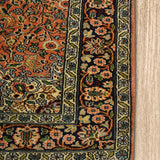 A close-up of the corner of a Kirman Persian rug, highlighting its intricate floral border design and delicate fringe. The rich peach and navy hues contrast beautifully with the finely detailed green and ivory patterns. The rug is placed on a light wooden floor, emphasizing its elegant craftsmanship.