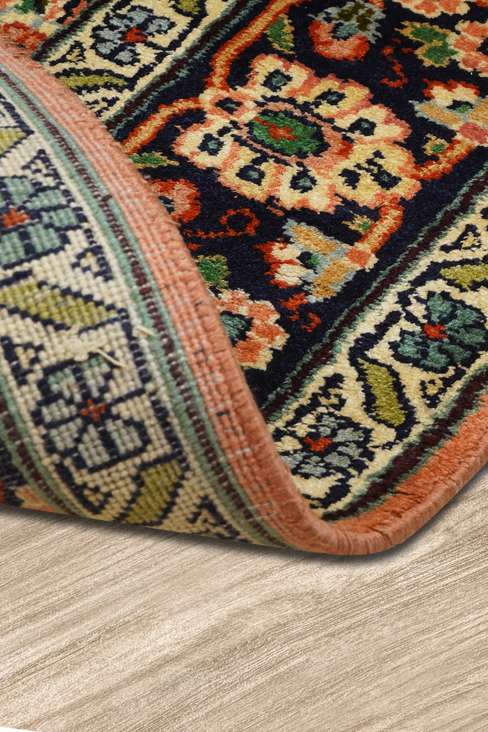 A close-up view of a Kirman Persian rug with a folded corner, showcasing its intricate floral border design and tightly woven backing. The rich navy, peach, and green hues highlight the traditional craftsmanship, while the rug's thickness and fine weave structure are clearly visible. The rug is placed on a light wooden floor, adding contrast to its ornate patterns.