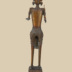 Back view of an antique dark brass sculpture of a tribal warrior, showing the details of the headdress, jewelry, and the way the sword and open hand are positioned. The texture of the brass and the decorated base are also visible.