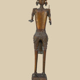 Back view of an antique dark brass sculpture of a tribal warrior, showing the details of the headdress, jewelry, and the way the sword and open hand are positioned. The texture of the brass and the decorated base are also visible.
