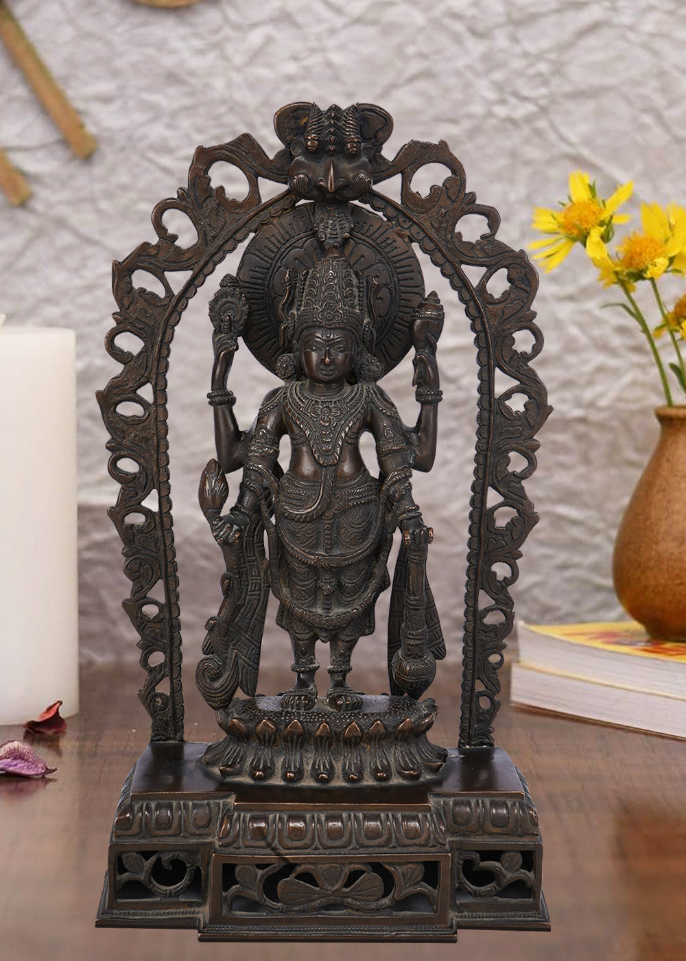 Dark brass statue of Lord Vishnu standing within an ornate arch, holding his traditional symbols: the conch shell, discus, lotus flower, and mace. The sculpture has a detailed finish and rests on a tiered pedestal.