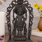 Dark brass statue of Lord Vishnu standing within an ornate arch, holding his traditional symbols: the conch shell, discus, lotus flower, and mace. The sculpture has a detailed finish and rests on a tiered pedestal.
