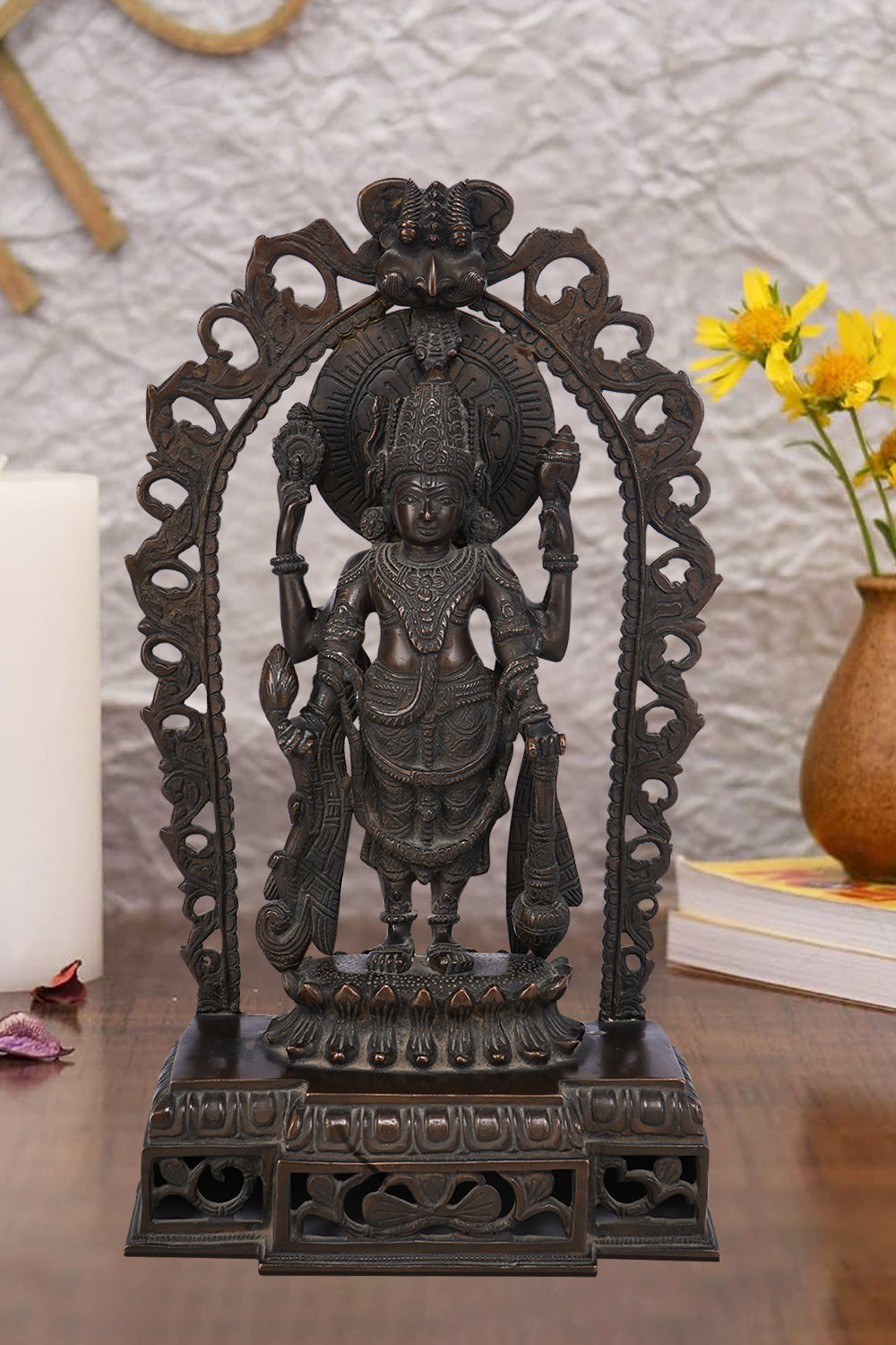 Dark brass statue of Lord Vishnu standing within an ornate arch, holding his traditional symbols: the conch shell, discus, lotus flower, and mace. The sculpture has a detailed finish and rests on a tiered pedestal.