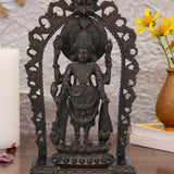 Dark brass statue of Lord Vishnu standing within an ornate arch, holding his traditional symbols: the conch shell, discus, lotus flower, and mace. The sculpture has a detailed finish and rests on a tiered pedestal.