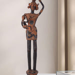 Dark brass figure depicting a rural woman carrying a small child in one arm and balancing a pot on her head. The antique-style sculpture features traditional adornments and stands on an ornate base.