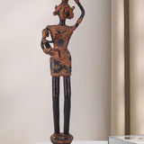 Dark brass figure depicting a rural woman carrying a small child in one arm and balancing a pot on her head. The antique-style sculpture features traditional adornments and stands on an ornate base.