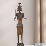 Dark brass figure of a tribal warrior holding a sword in one hand and an open palm in the other, featuring intricate jewelry, a unique headdress, and standing on a decorated pedestal.