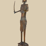 Vintage-style dark brass sculpture of a warrior holding a curved sword and with an open palm, adorned with traditional attire and a unique headdress, standing on a decorated platform.
